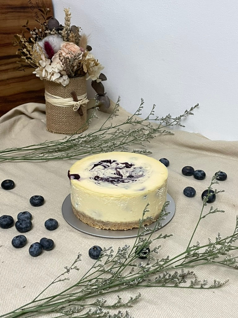 Cheesecake with blueberries on a table with flowers and a vase in the background